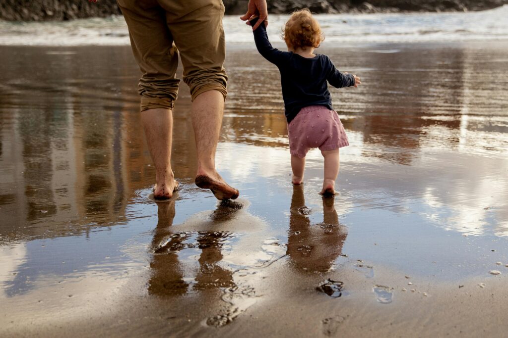 A father and toddler enjoy a sunny day walking barefoot on a sandy beach, capturing a moment of family bonding.