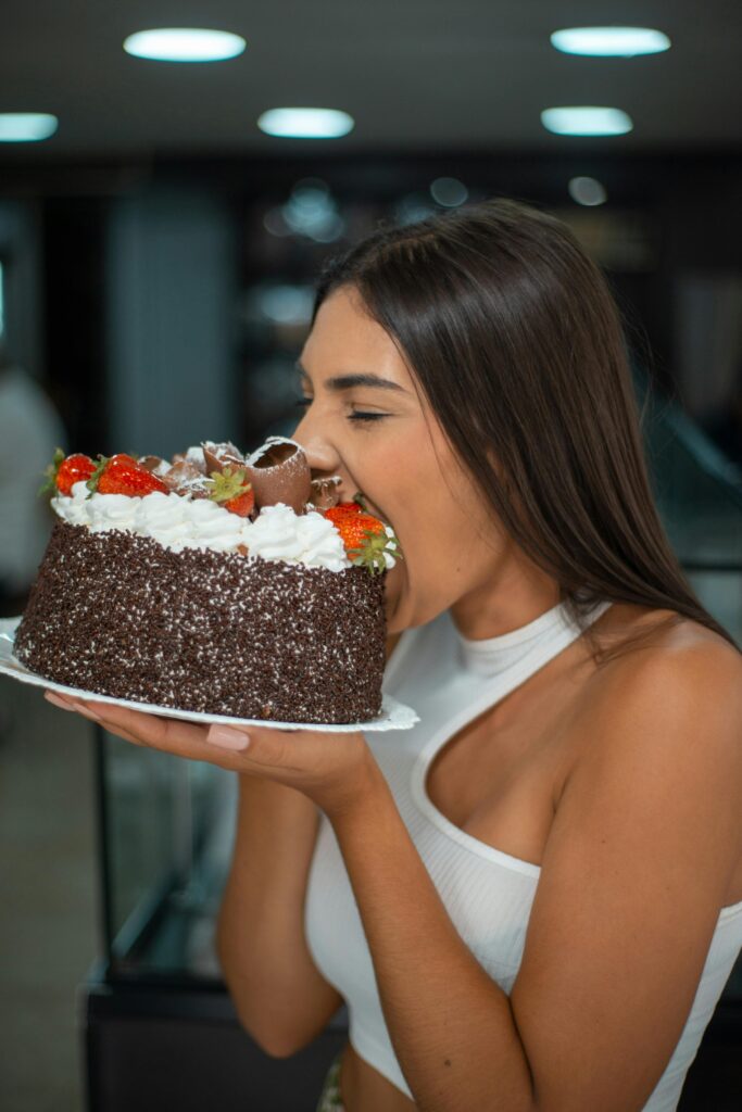 A young brunette woman stress eating, bites into a chocolate frosting cake topped with strawberries and white frosting indoors.